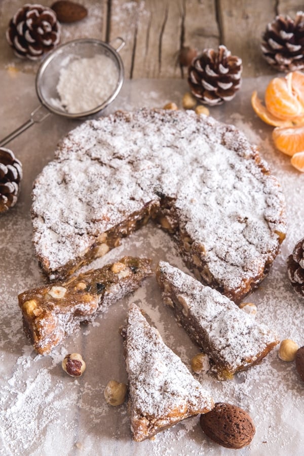 Round fruit and nut cake dusted with powdered sugar, with several slices cut, surrounded by pinecones and nuts—an inviting centerpiece inspired by classic Italian dessert recipes.