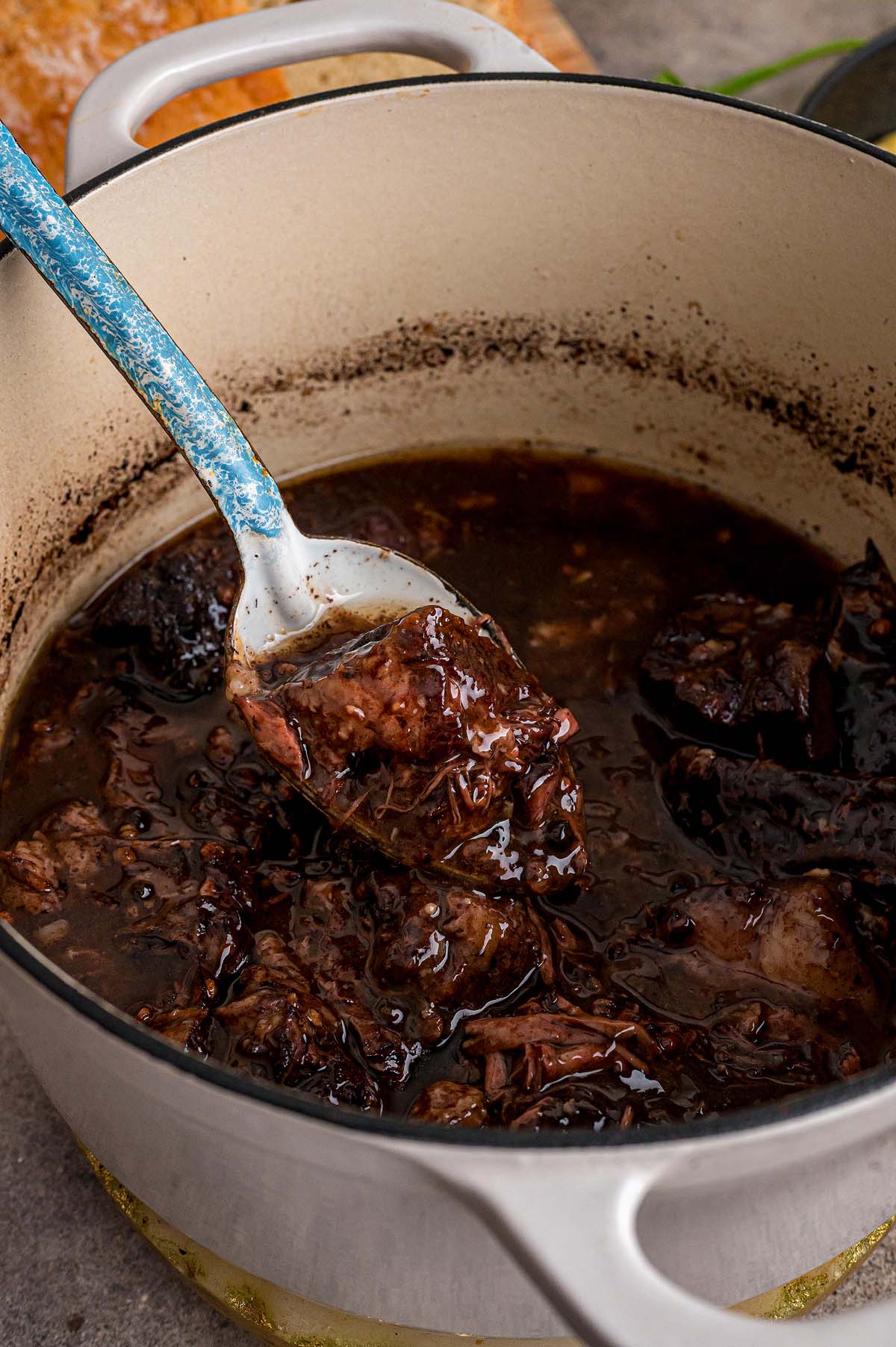 A blue spoon lifts shredded beef in dark peposo sauce from a white pot.