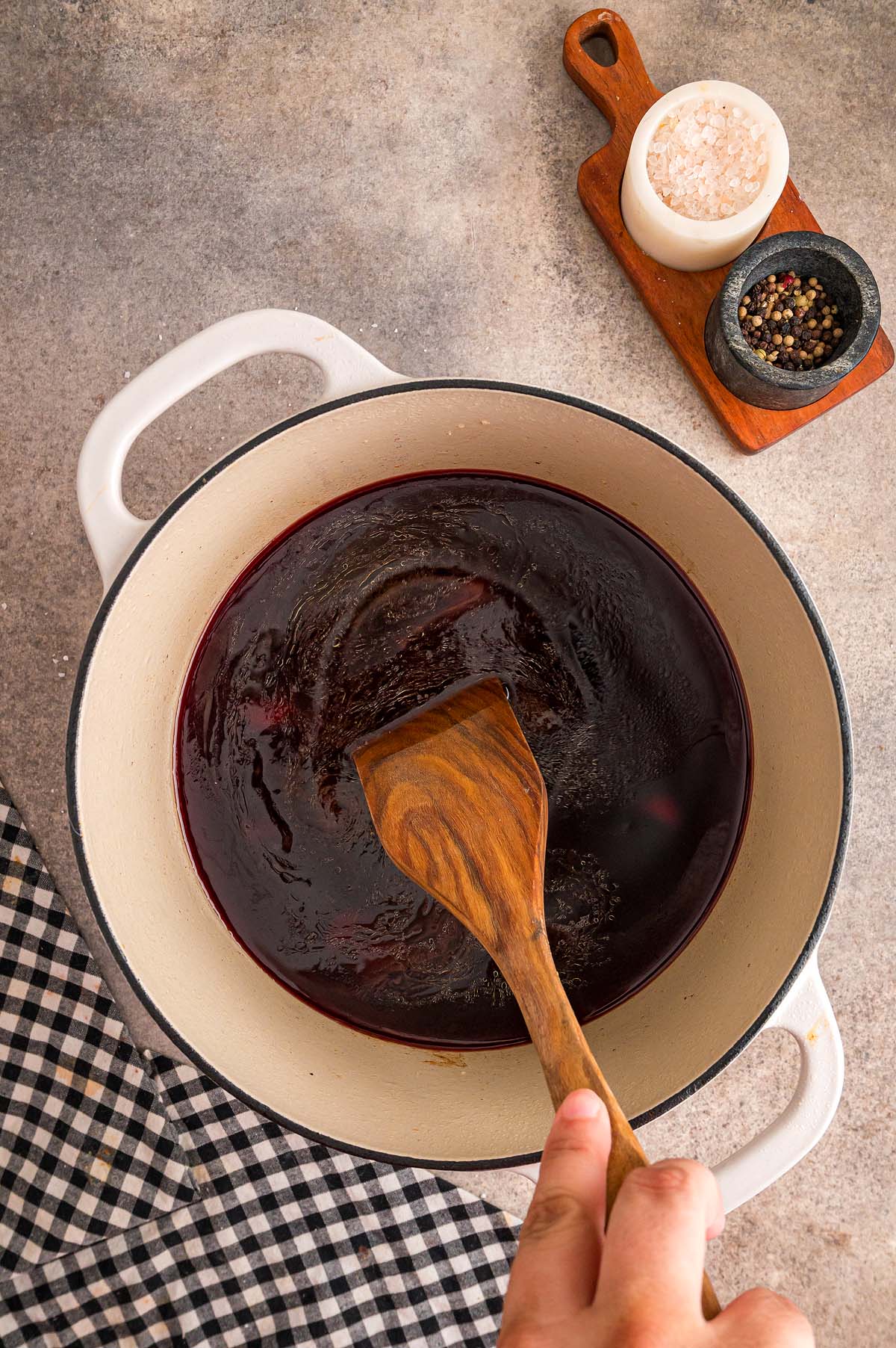 A hand stirs dark liquid in a white pot with a wooden spatula; salt and pepper bowls nearby, hinting at a hearty peposo in the making.