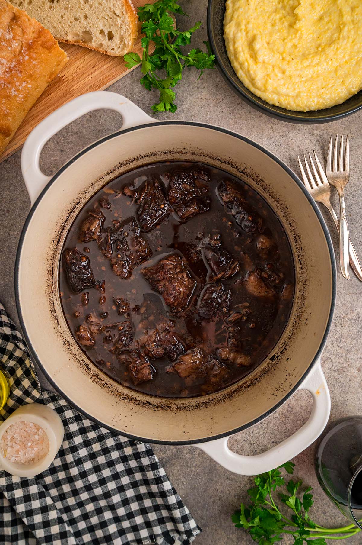 A white pot of braised short ribs in a rich peposo sauce sits on the table, surrounded by polenta, bread, herbs, and forks.
