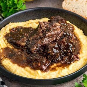 Peposo served on creamy polenta in a black bowl, with bread and herbs on the side.