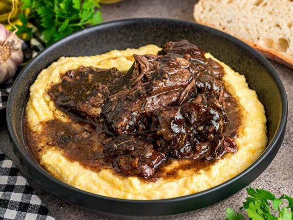 Peposo served on creamy polenta in a black bowl, with bread and herbs on the side.