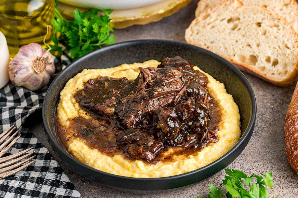 Peposo beef stew served over creamy polenta in a black bowl, with bread, garlic, and herbs on the side.