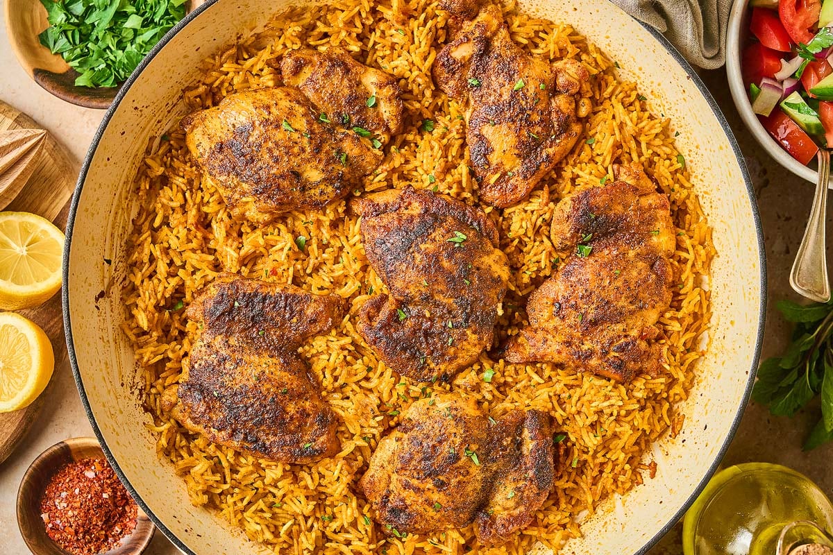 A Shawarma Chicken and Rice Skillet features seasoned chicken thighs on spiced rice, surrounded by lemon slices, fresh herbs, and a crisp salad.