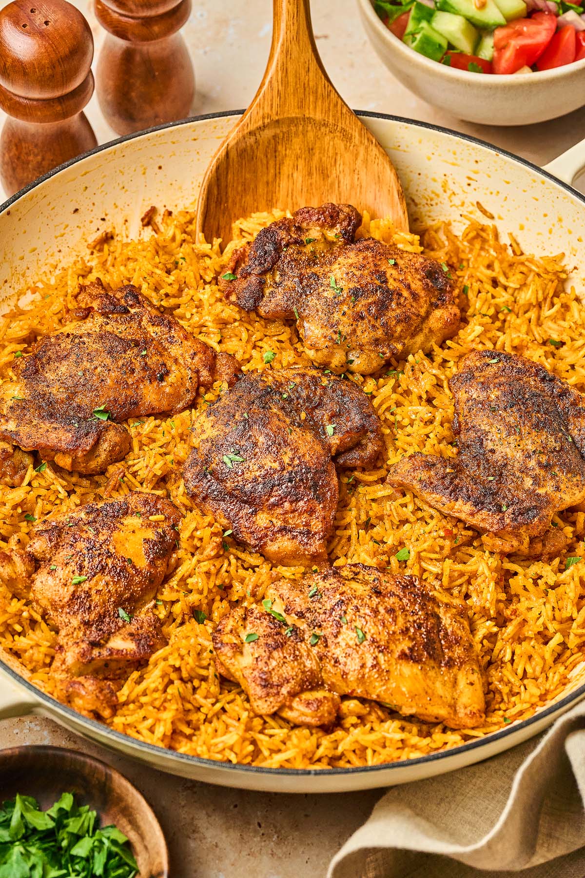 A Shawarma Chicken and Rice Skillet features seasoned chicken thighs on yellow rice, with a wooden spoon and a bowl of salad nearby.