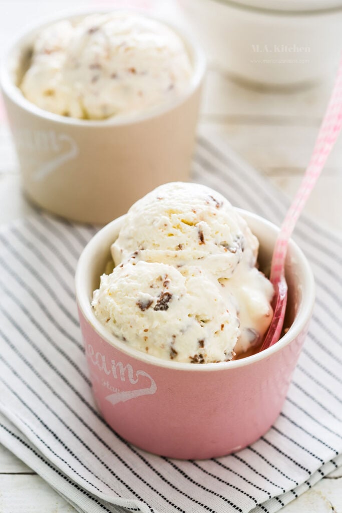 Two scoops of vanilla ice cream with chocolate chips in a pink cup with a spoon, inspired by classic Italian dessert recipes, served on a striped cloth.