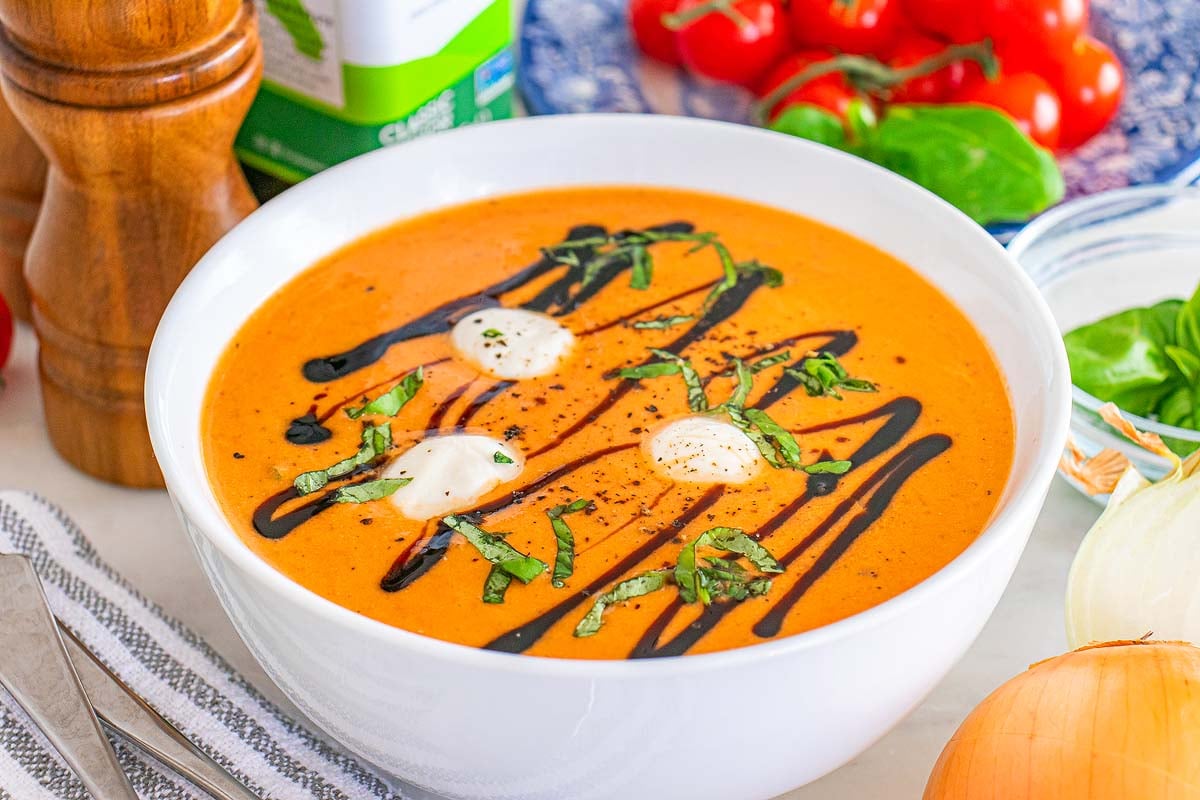 Bowl of tomato Caprese soup topped with cream, basil, and balsamic glaze, surrounded by fresh ingredients.