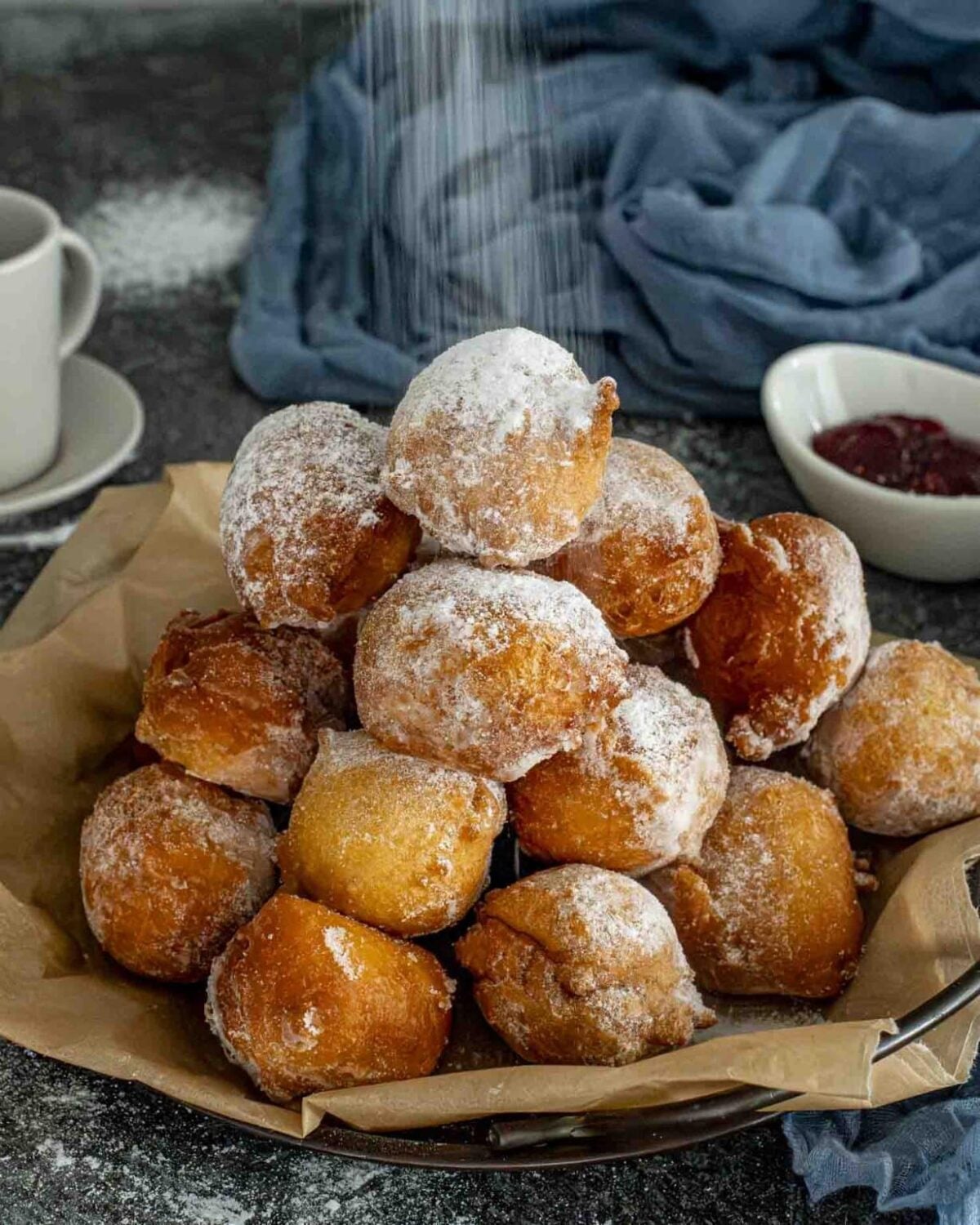 A pile of powdered sugar-covered fried doughnuts on parchment paper, inspired by Italian dessert recipes, with coffee and jam in the background.