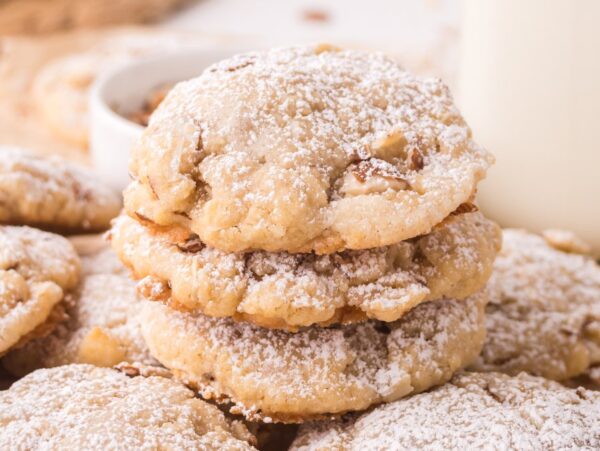 A stack of Almond Croissant Cookies, dusted with powdered sugar and filled with nuts, sits next to a glass of milk on the table.