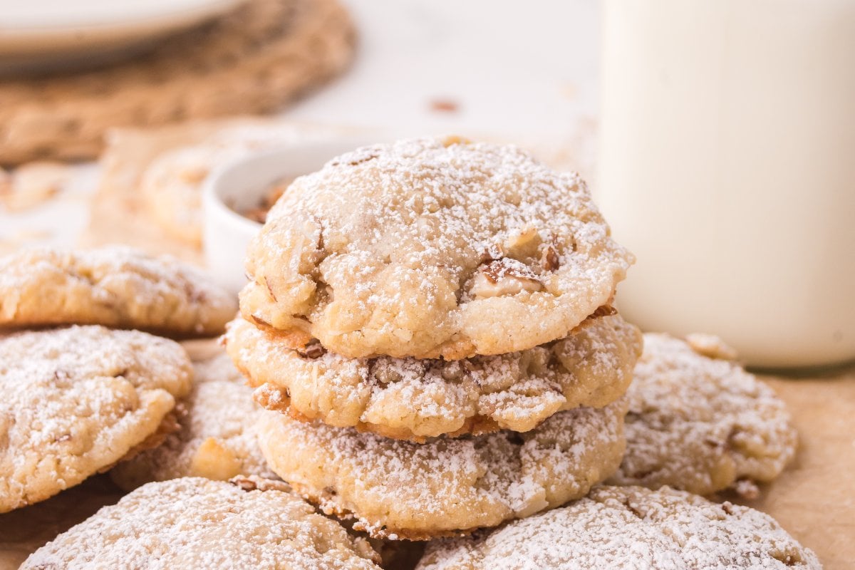 A stack of powdered sugar-dusted Almond Croissant Cookies sits beside a glass of milk on a light surface.