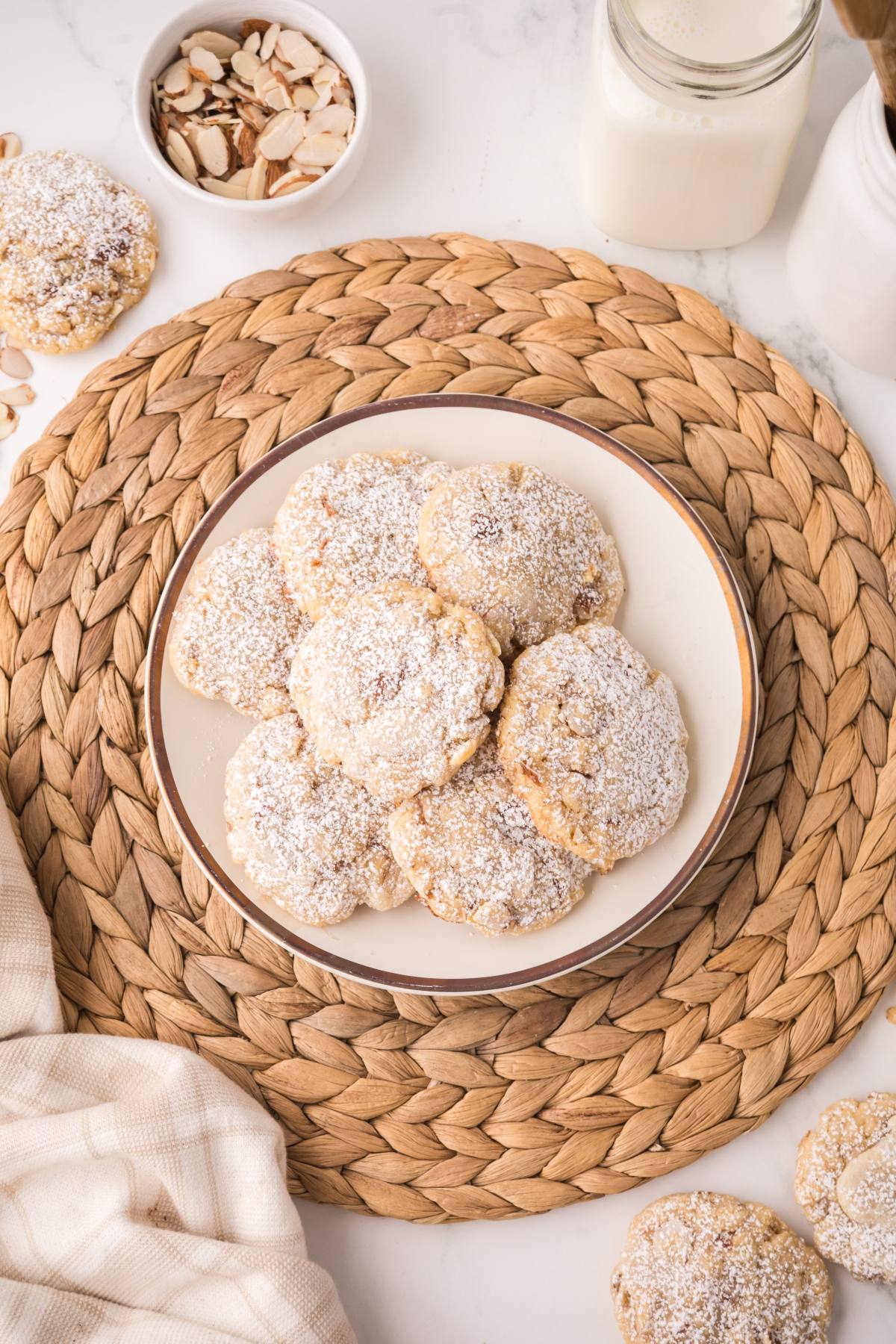 A plate of powdered sugar almond cookies on a woven placemat, with sliced almonds and a jar of milk nearby.
