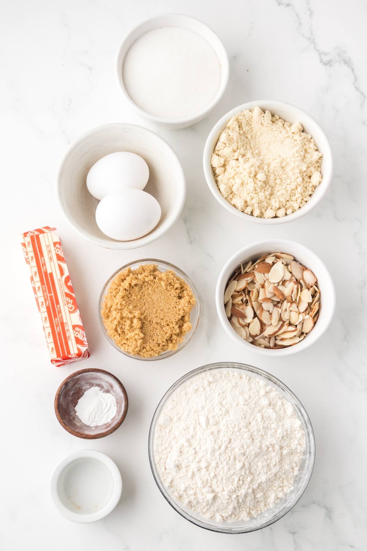 Baking ingredients on a white surface: eggs, sugar, flour, butter, brown sugar, sliced almonds, and baking powder.