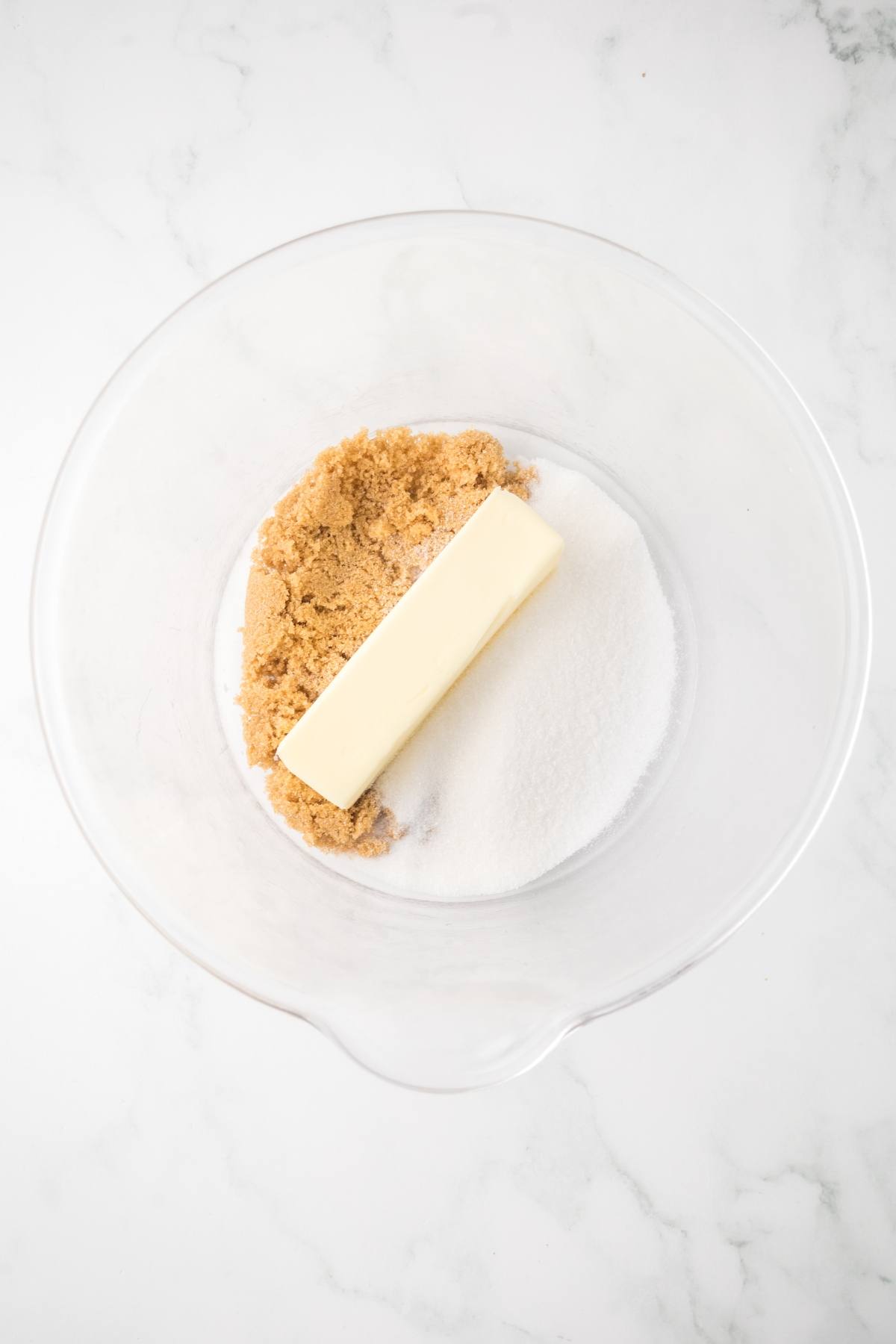 A clear bowl with a stick of butter, brown sugar, and white sugar on a white marble surface.