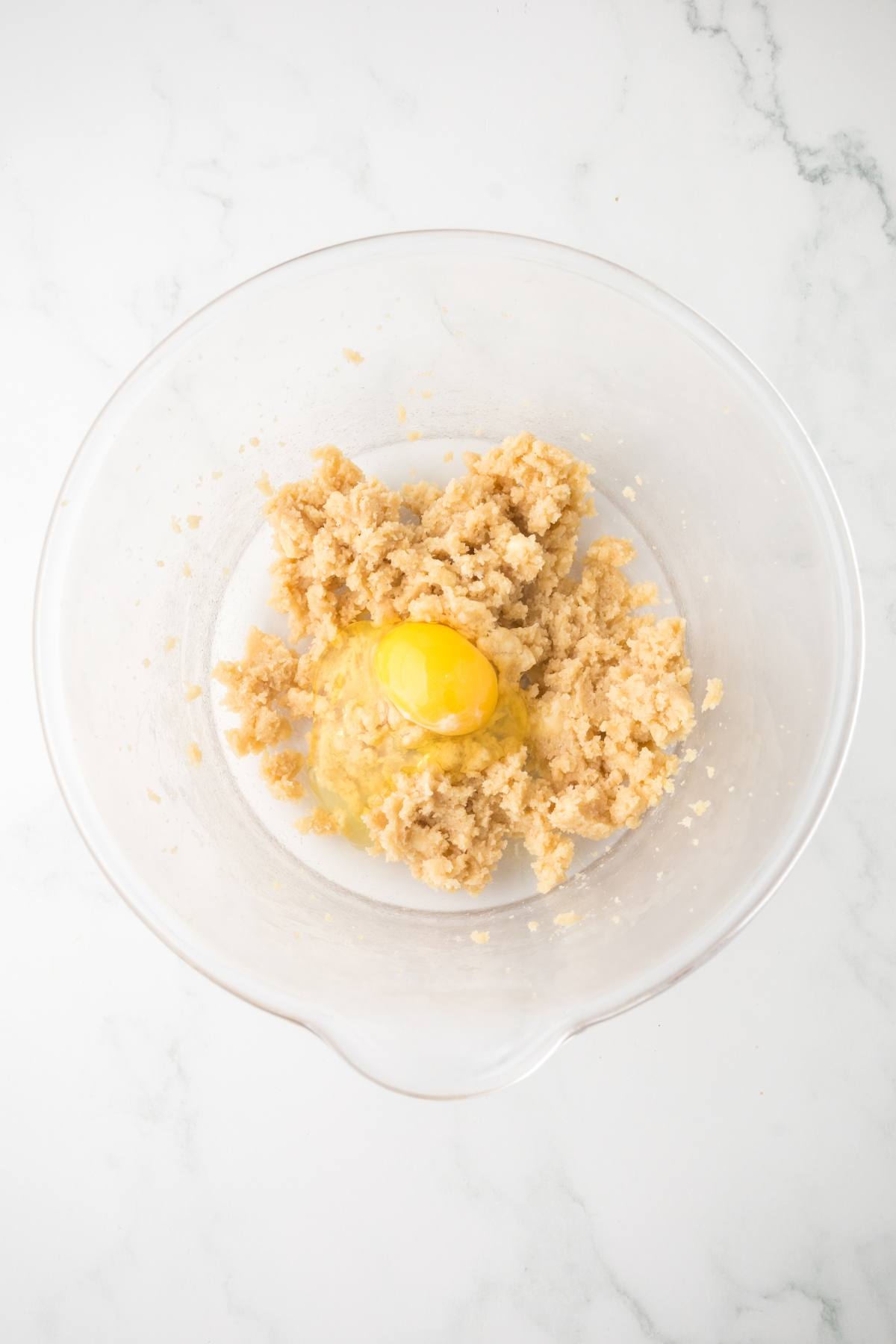 A glass mixing bowl with cookie dough and a cracked raw egg on a white marble surface.