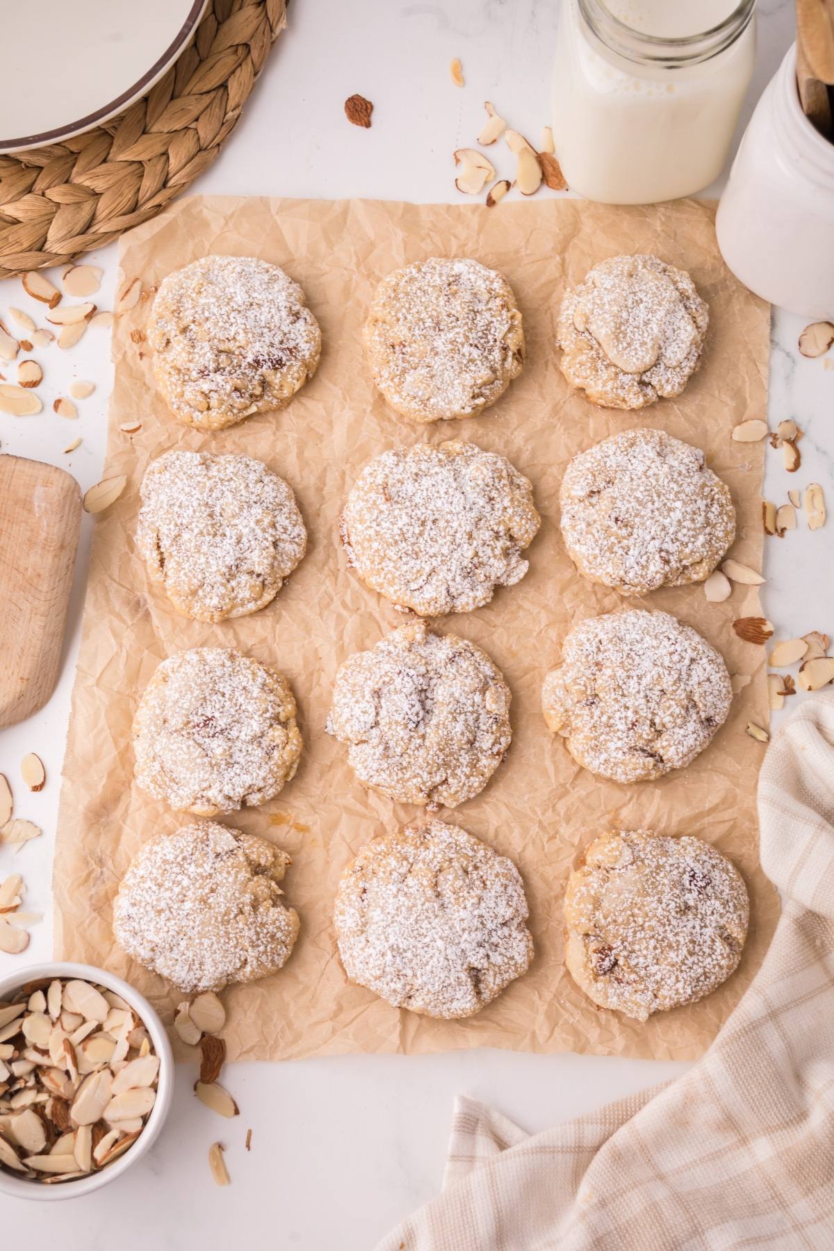 Twelve powdered sugar cookies on parchment paper with sliced almonds and a glass of milk nearby.