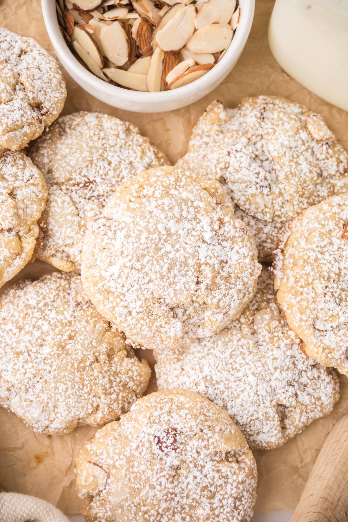 Powdered sugar-covered cookies on parchment paper, with a bowl of sliced almonds in the background.