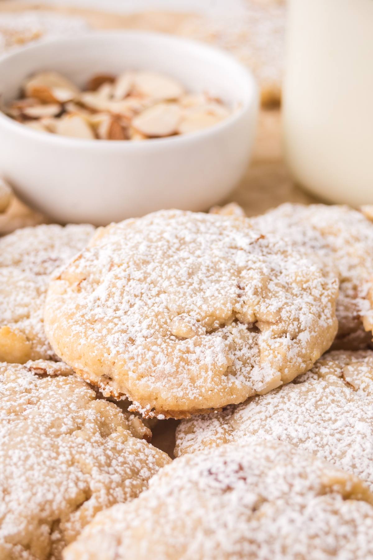 Close-up of almond cookies dusted with powdered sugar, with a bowl of sliced almonds in the background.