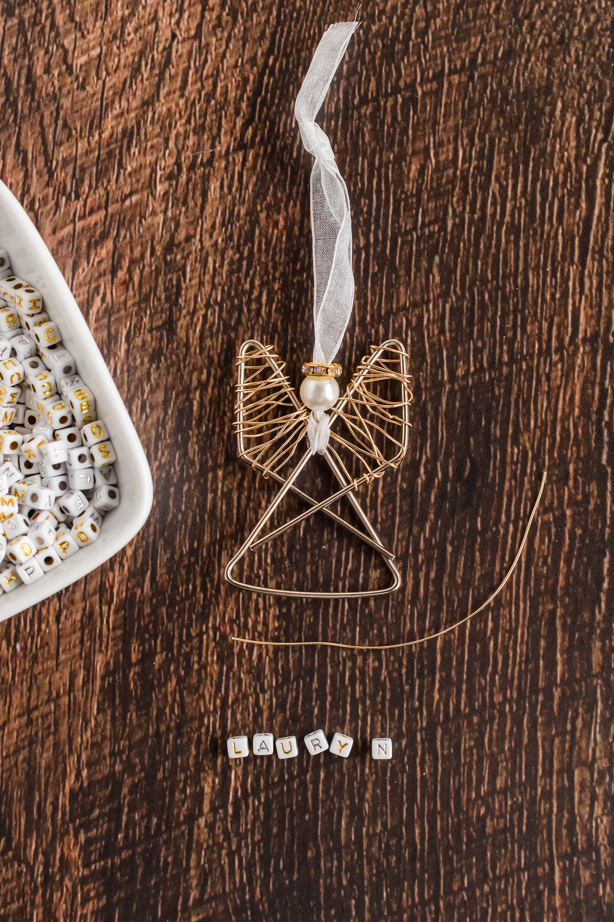 Gold wire angel ornament with "Lauryn" beads beside a dish of alphabet beads on a wooden surface—an easy personalized paper clip angel craft idea.