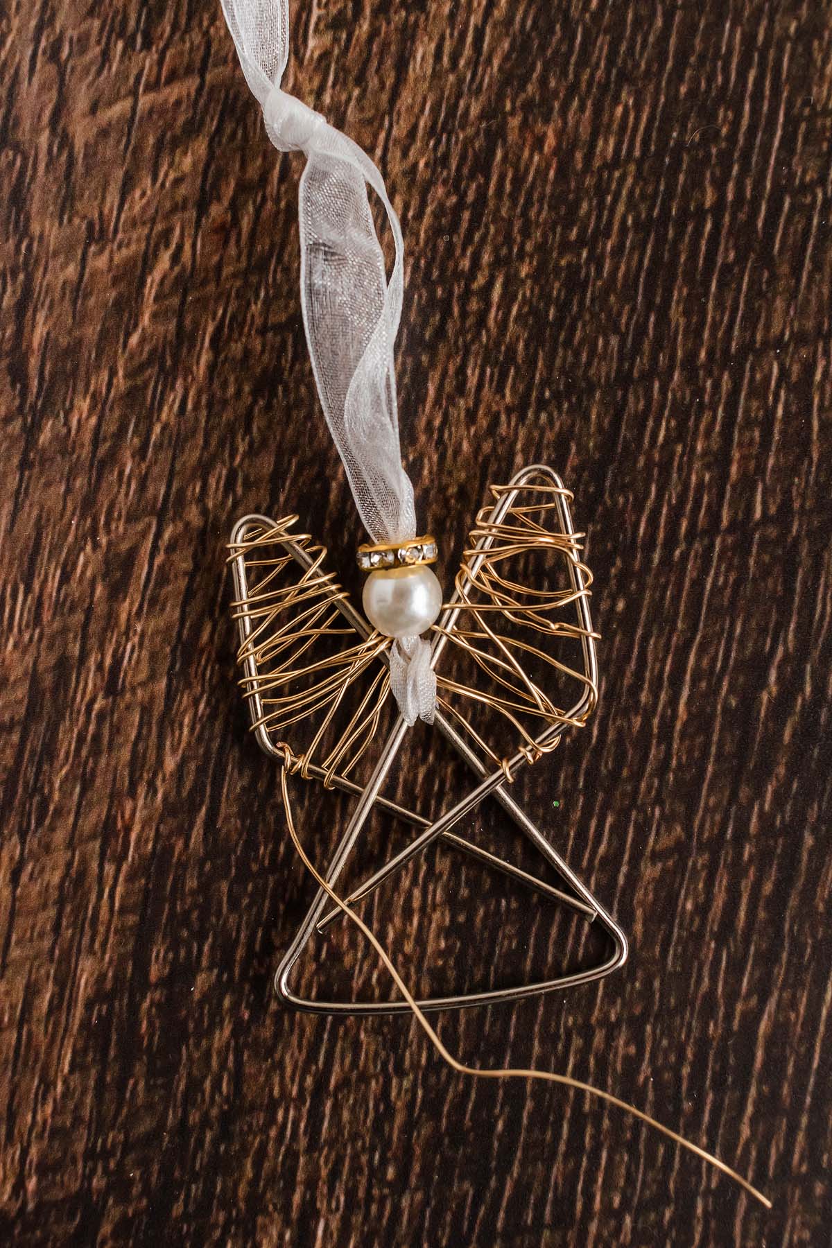 Easy personalized paper clip angel ornament featuring a pearl bead head and gold halo, tied with a sheer ribbon, displayed on a wooden surface.