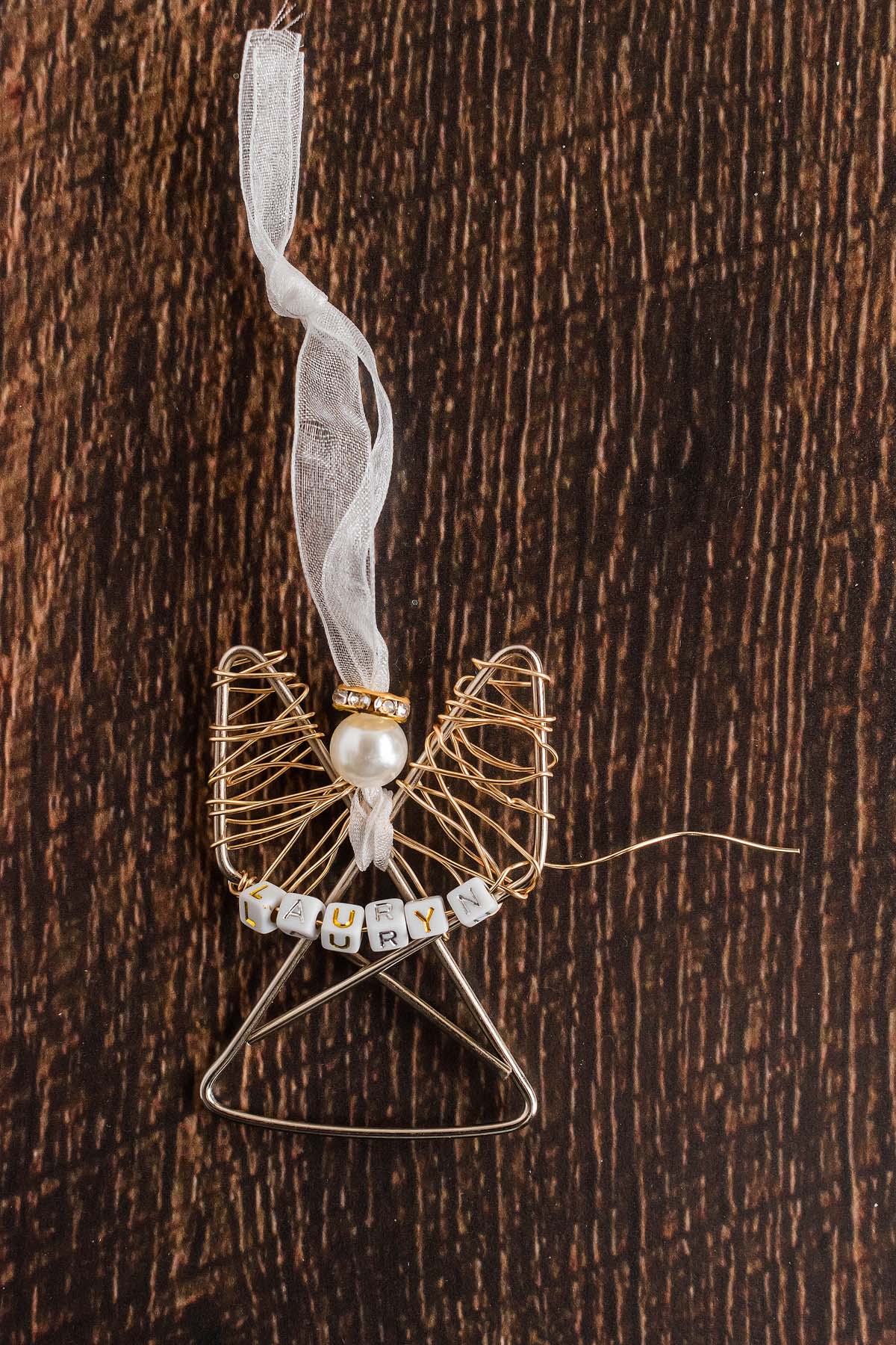 Easy personalized paper clip angel ornament with a ribbon, pearl bead, and letter beads spelling "LAURYN," displayed on a wooden background.