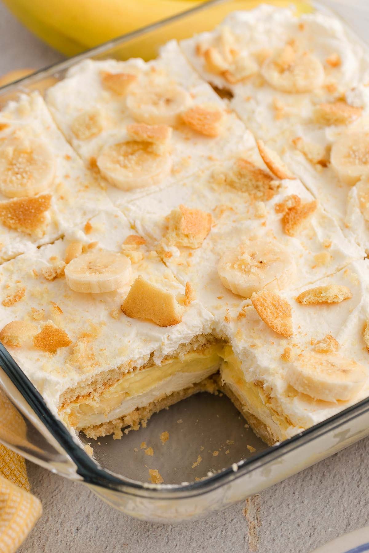 Glass dish of classic Banana Pudding with whipped topping, vanilla wafers, and banana slices; one creamy serving removed.