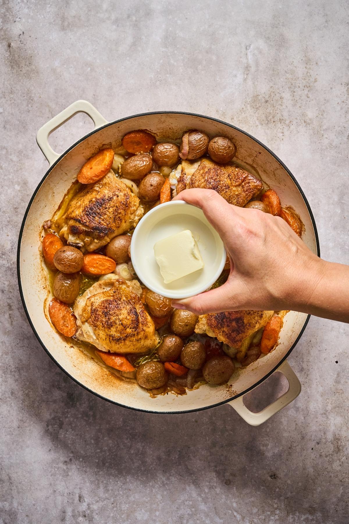 A hand adds a bowl of butter to a pot of roasted chicken, carrots, and potatoes.