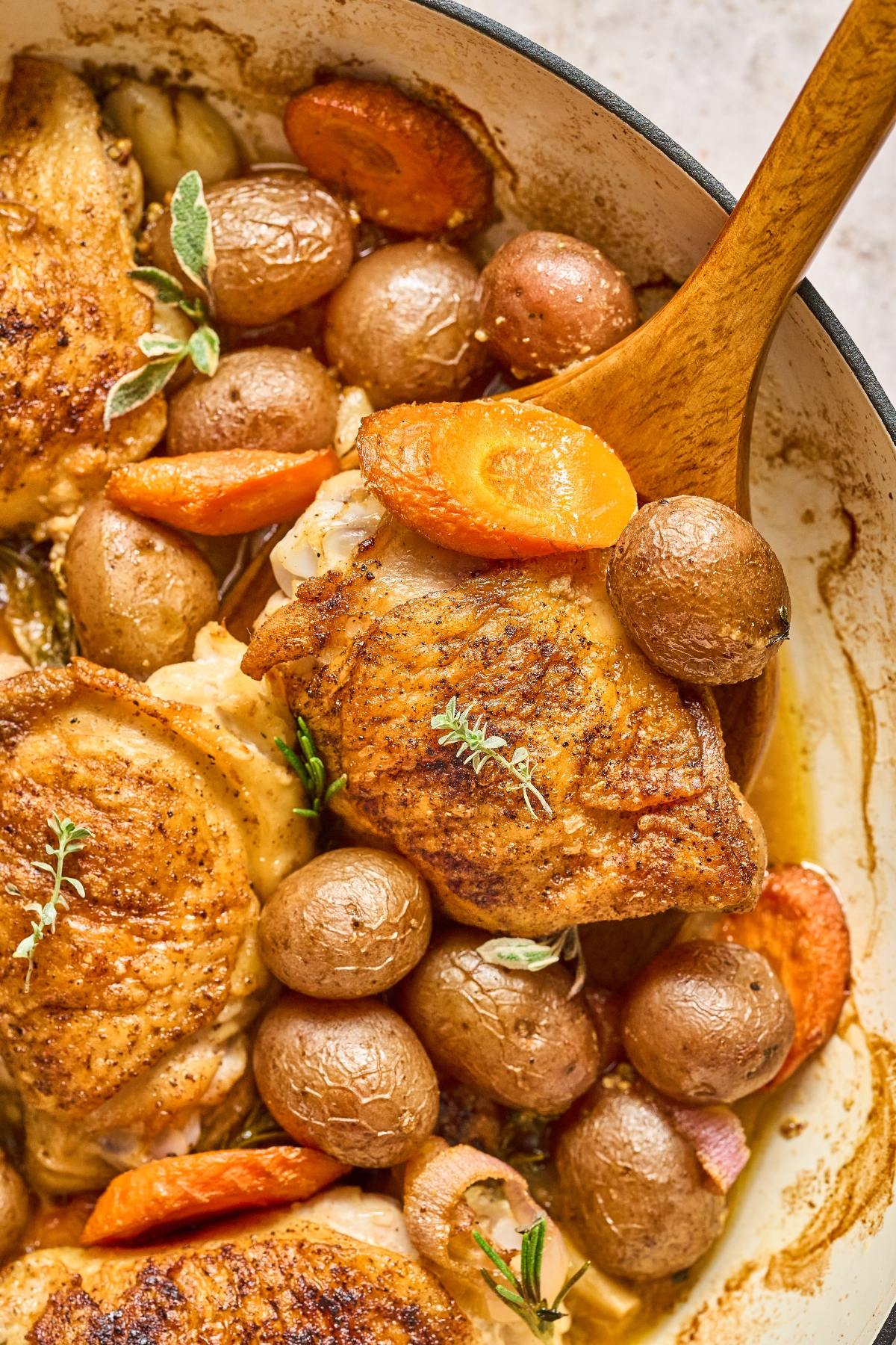 Golden roasted chicken thighs with baby potatoes, carrots, and herbs in a pan, served with a wooden spoon.