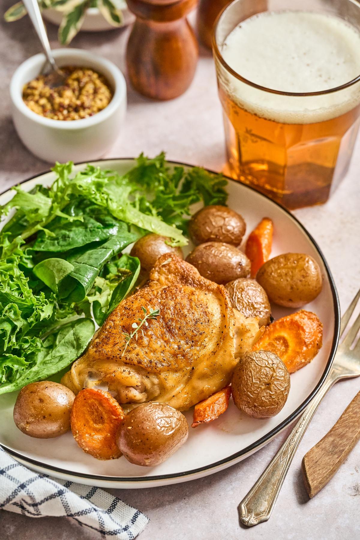 Roast chicken with baby potatoes, carrots, and greens on a plate next to a glass of beer.