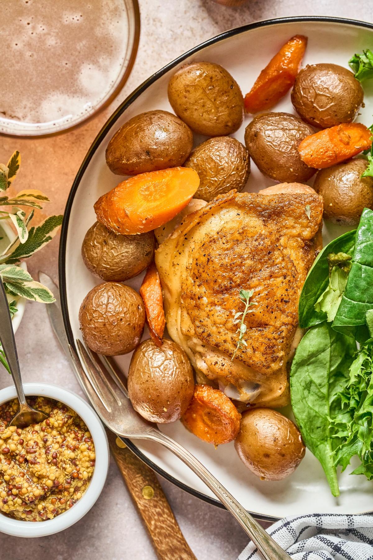 Roast chicken thigh with baby potatoes, carrots, greens, and mustard on a plate with a drink nearby.