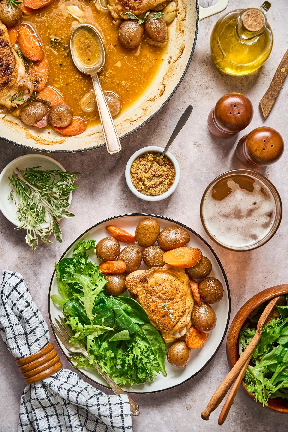 A plate with roasted chicken, potatoes, carrots, and salad, with mustard, herbs, and a drink on the table.