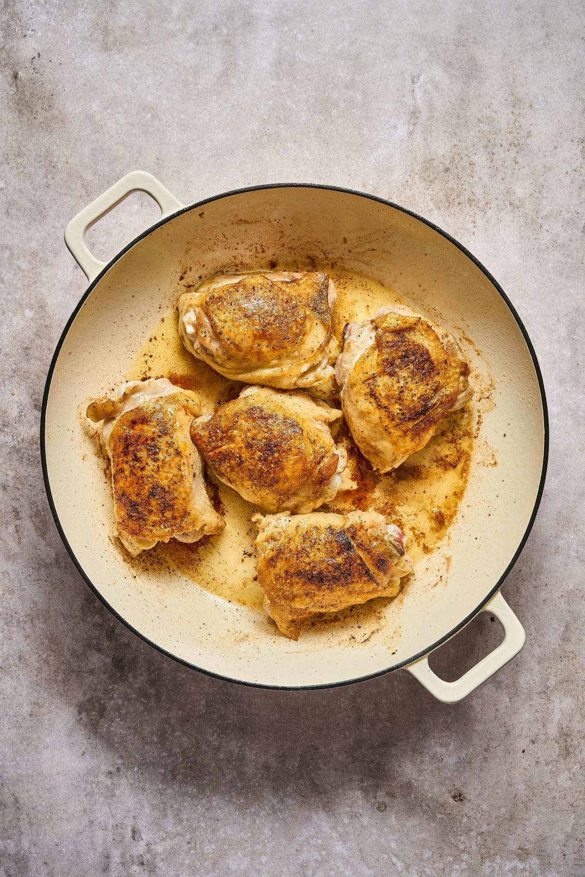 Five browned chicken thighs in a cream-colored skillet on a light gray textured surface.