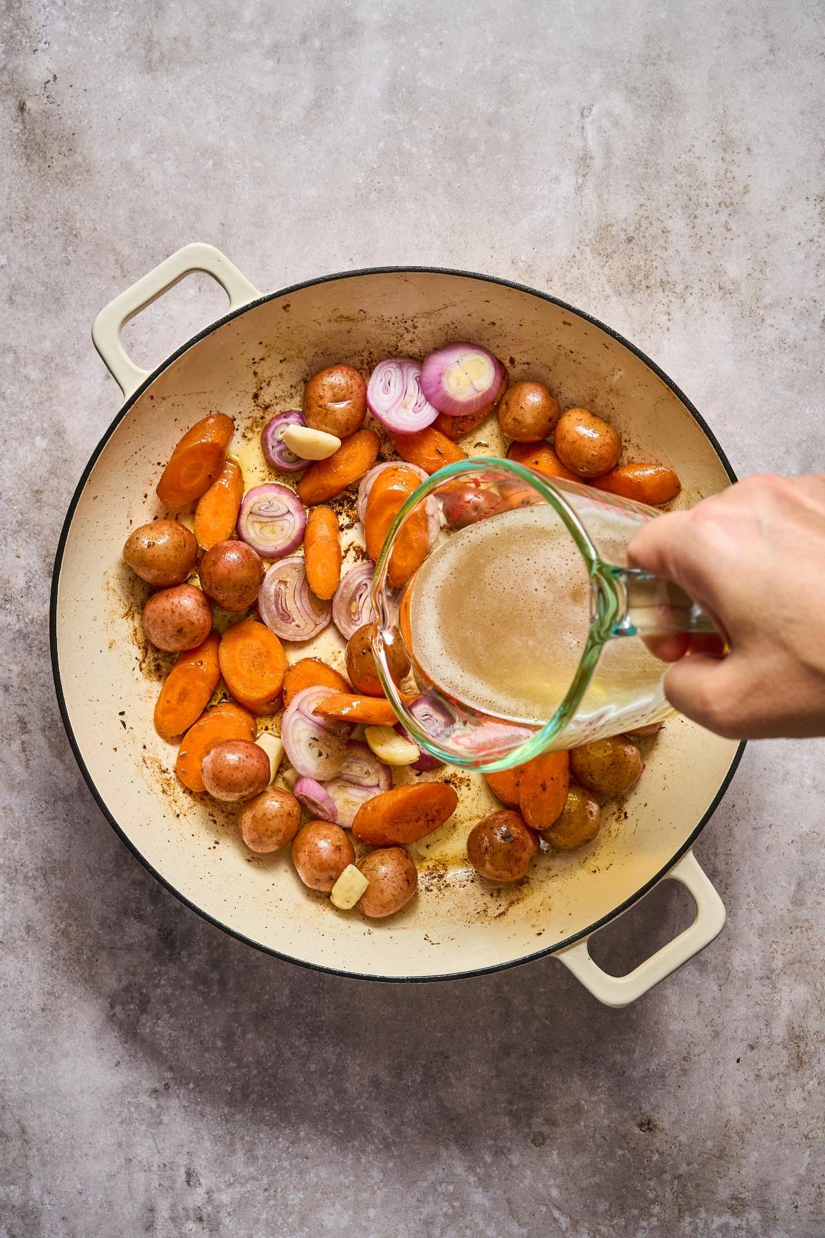A hand pours broth into a pot with potatoes, carrots, and sliced shallots on a light surface.