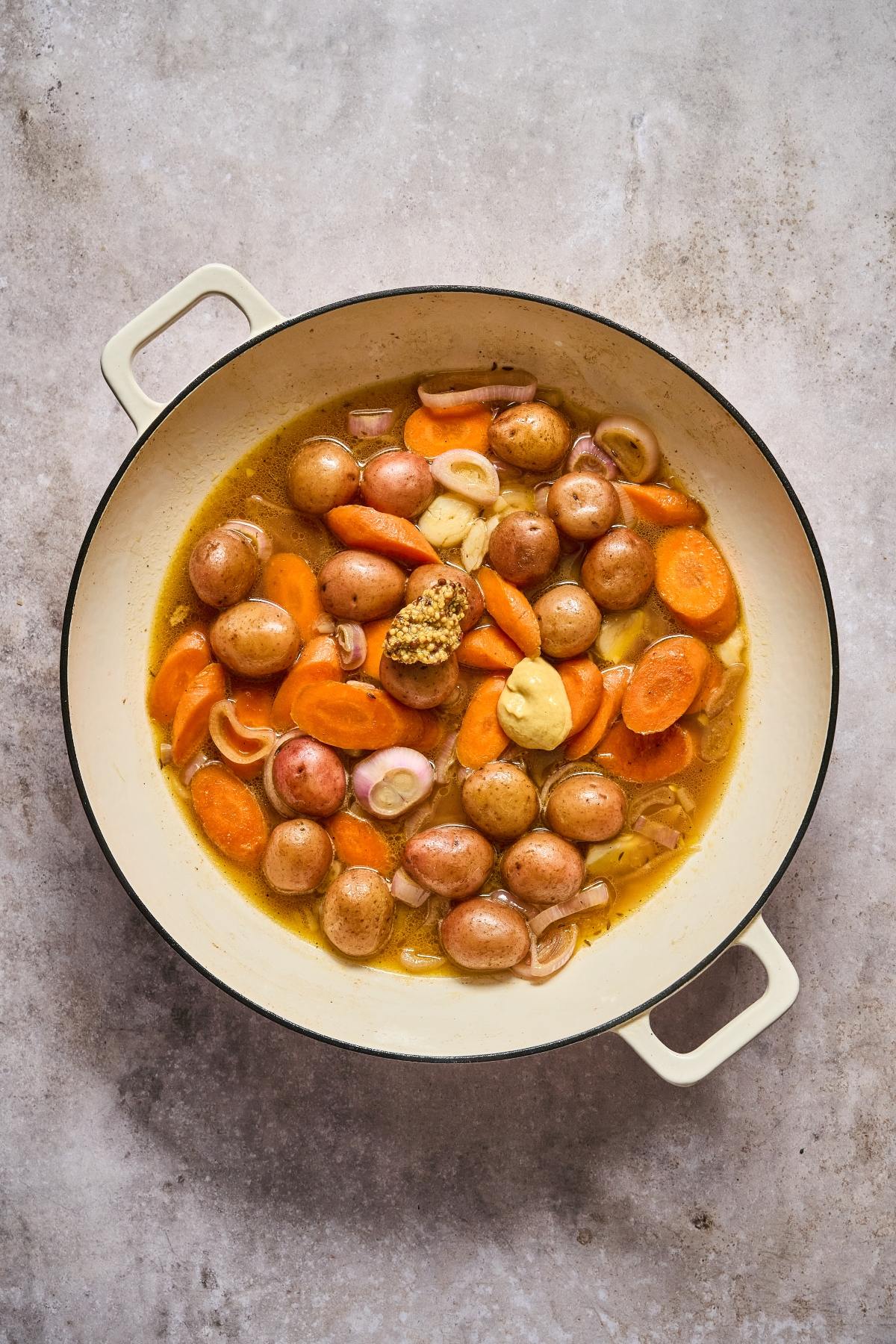 Round pot filled with baby potatoes, sliced carrots, shallots, and mustard in a broth, on a light surface.