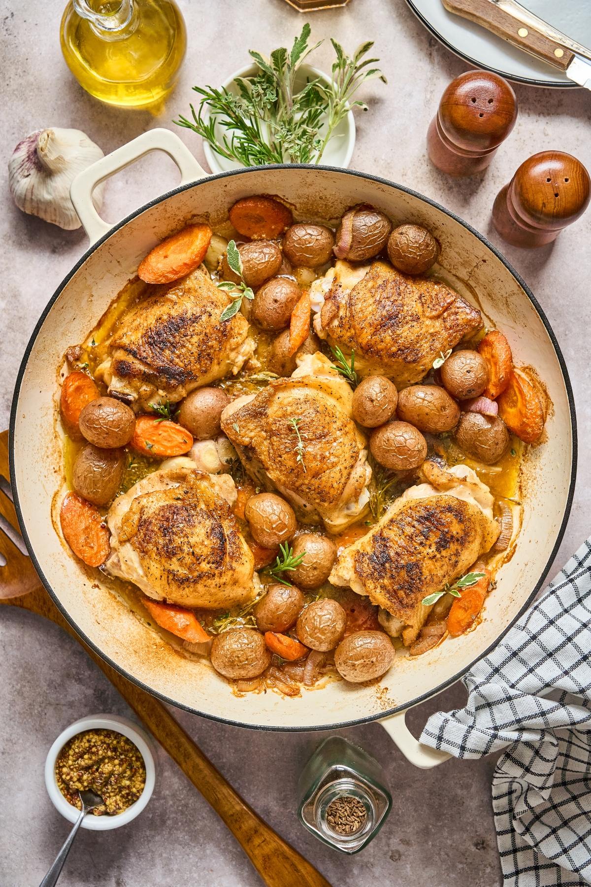 Roast chicken thighs with carrots and baby potatoes in a white pan, surrounded by herbs and kitchen utensils.