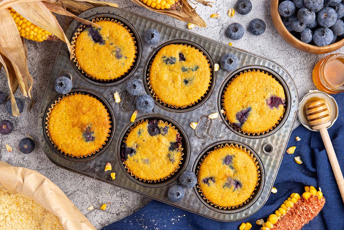 A muffin tin with six Blueberry Cornbread Muffins, surrounded by fresh blueberries, corn, and a honey dipper.