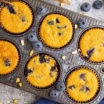 Six Blueberry Cornbread Muffins sit in a baking tray, surrounded by fresh blueberries, honey, corn, and baking ingredients.