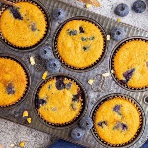 Six Blueberry Cornbread Muffins sit in a baking tray, surrounded by fresh blueberries, honey, corn, and baking ingredients.