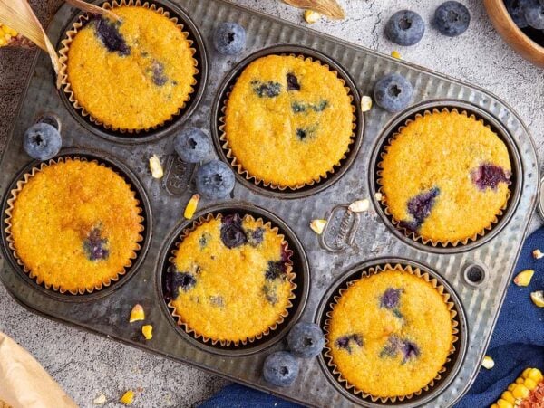Six Blueberry Cornbread Muffins sit in a baking tray, surrounded by fresh blueberries, honey, corn, and baking ingredients.