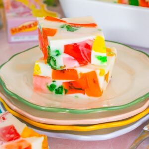 Three colorful Broken Glass Jello dessert squares on stacked pastel plates, with cutlery and more gelatin in the background.