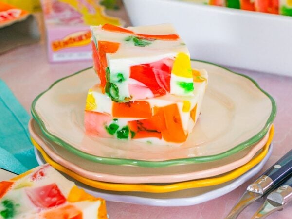 Three colorful Broken Glass Jello dessert squares on stacked pastel plates, with cutlery and more gelatin in the background.