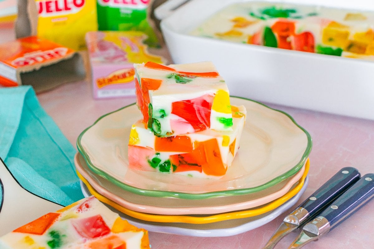 Three colorful Broken Glass Jello cubes with a creamy white layer are stacked on plates, with utensils and boxes in the background.