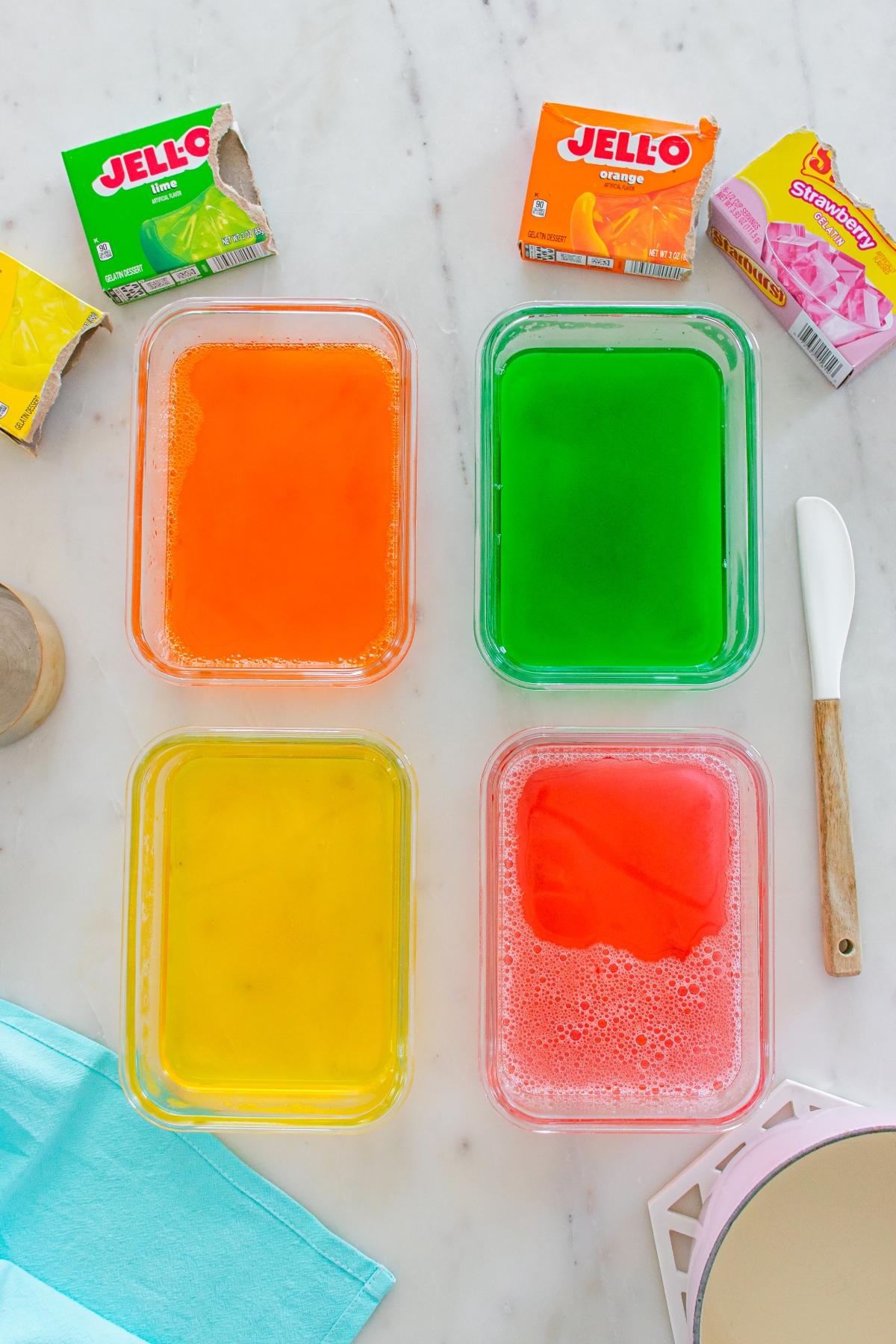 Four containers of colorful Jello&mdash;orange, green, yellow, and red&mdash;on a white countertop with Jello boxes nearby.