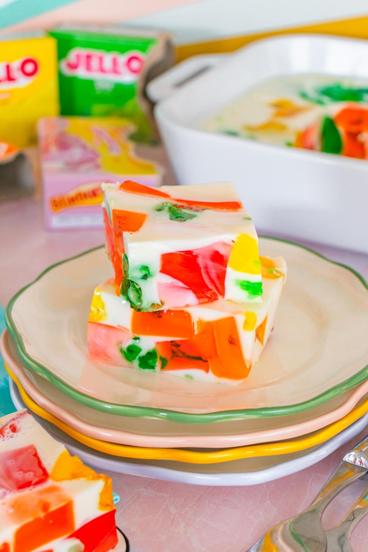 Three colorful Jello mosaic dessert squares stacked on a plate, with Jello boxes and a dish in the background.