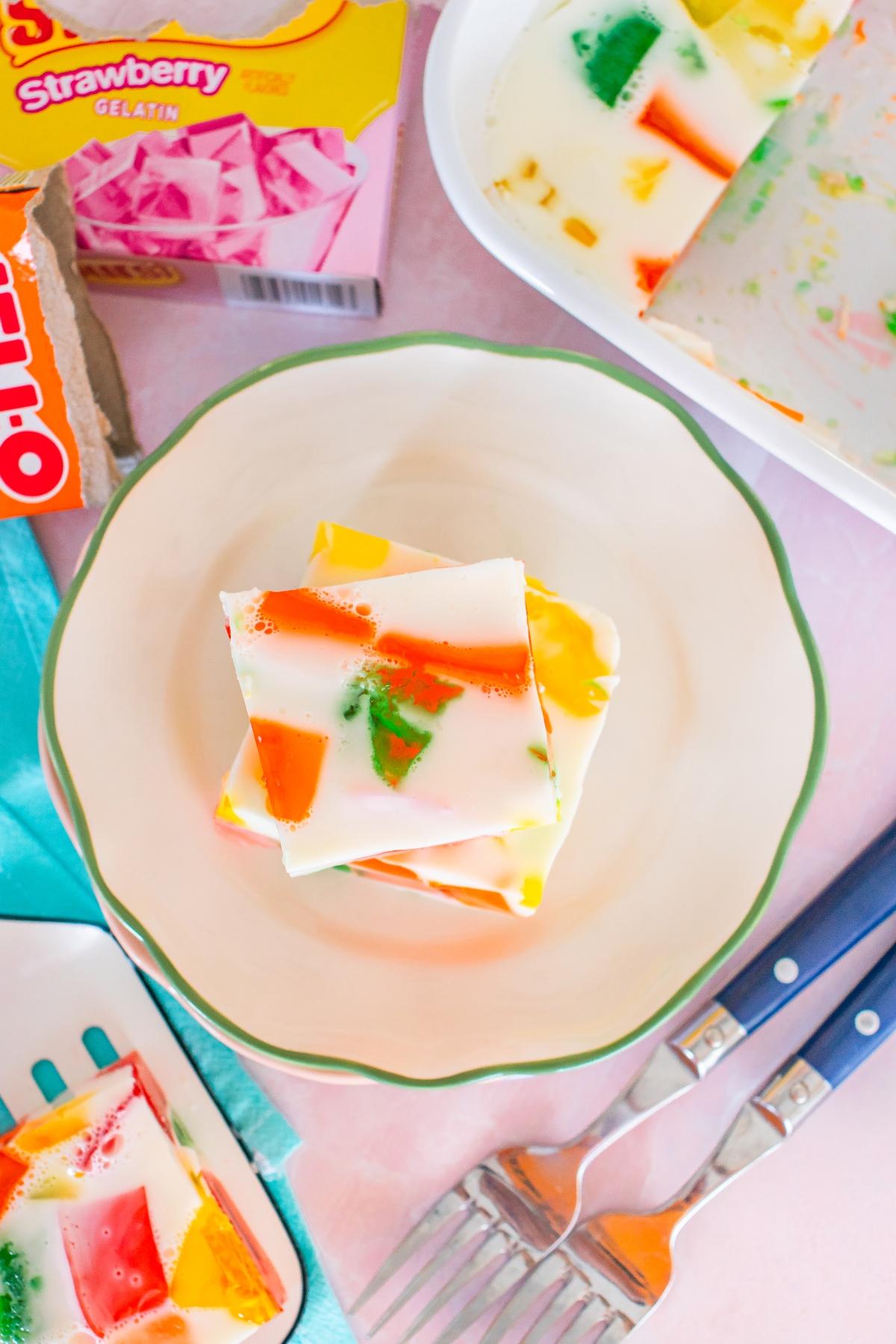 A plate with colorful Jello salad squares, surrounded by utensils and boxes of gelatin mix.