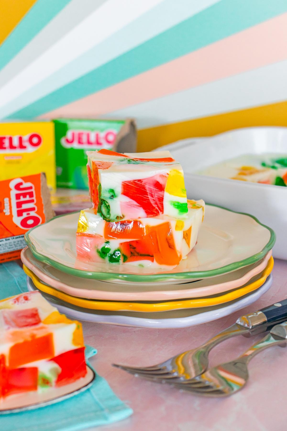Colorful layered Jello squares with creamy white base on stacked plates, with Jello boxes and utensils nearby.