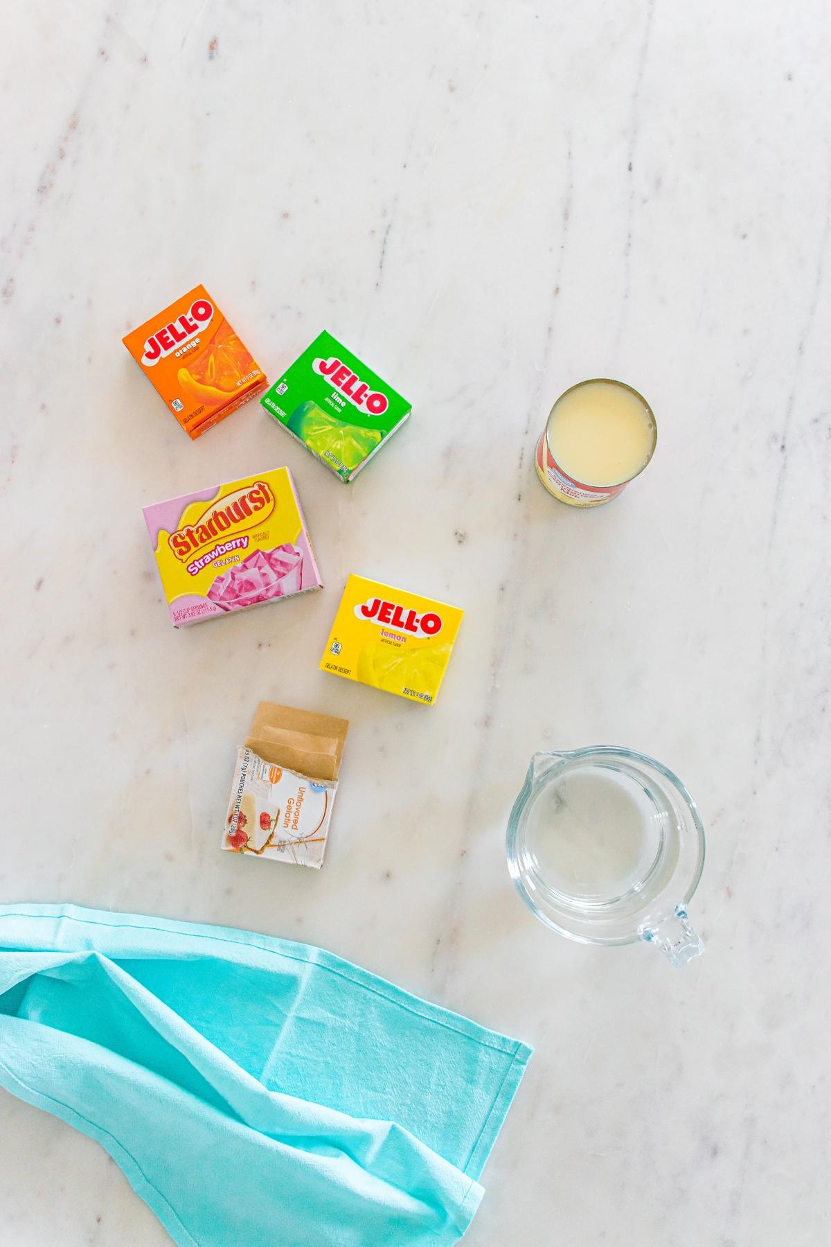 Jello boxes, Starburst pudding, condensed milk, a glass measuring cup, and a turquoise cloth on a marble surface.