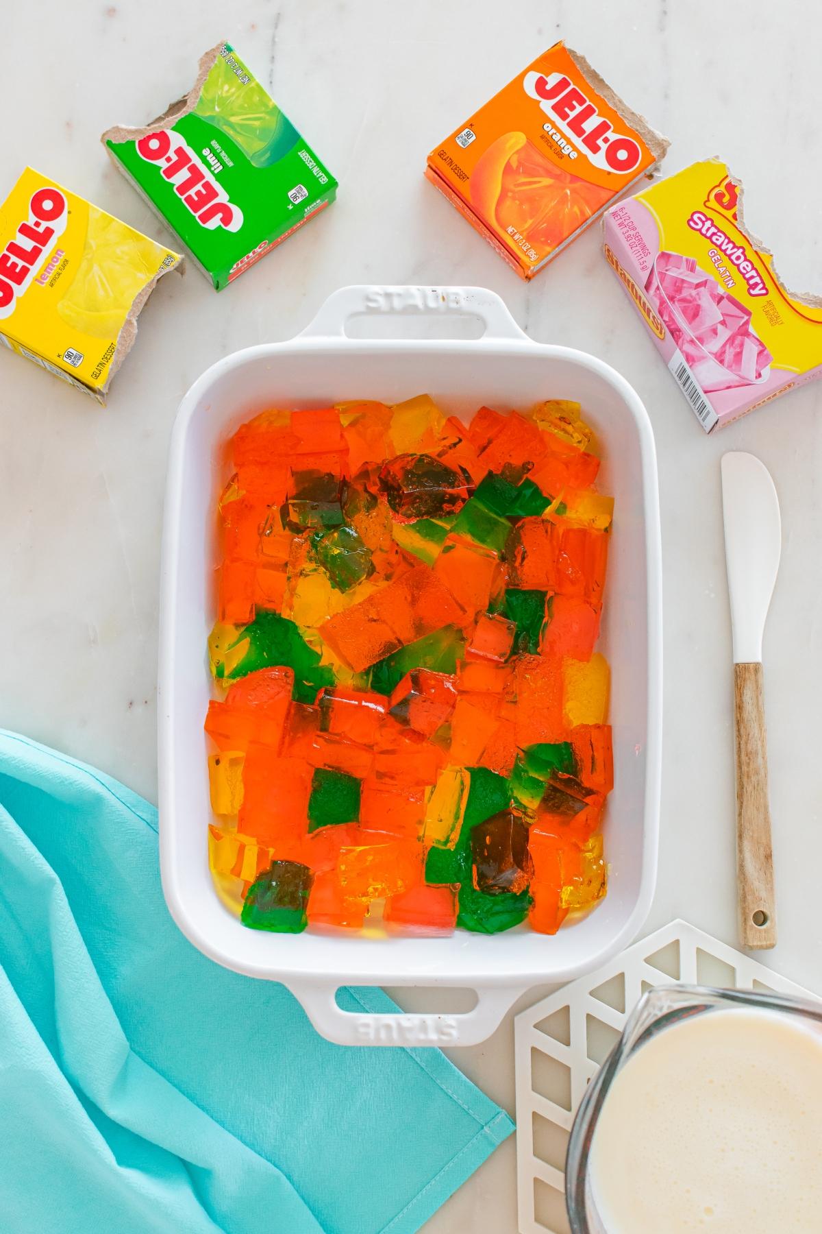 A white dish filled with colorful Jello cubes, Jello boxes, a spatula, and a blue cloth nearby.