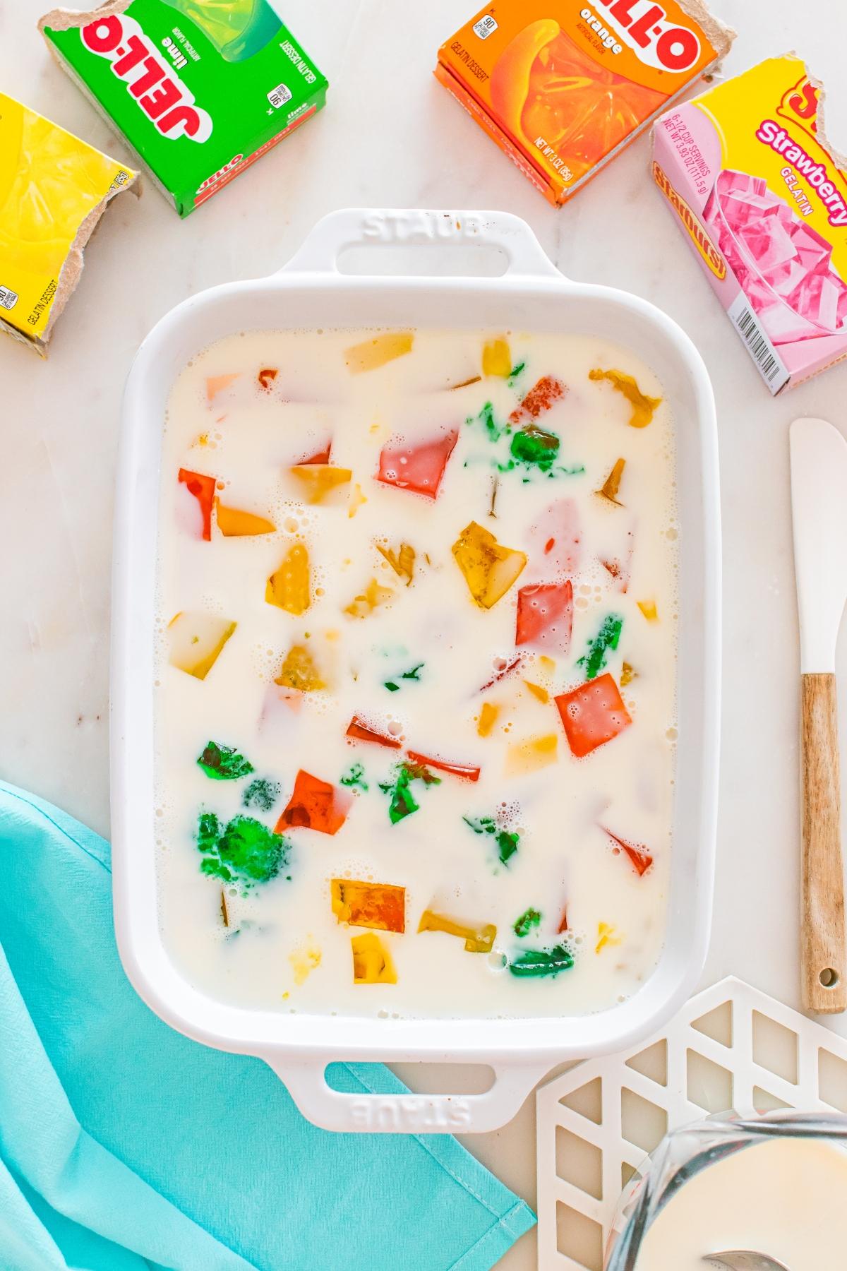 White dish with colorful Jello cubes in milky liquid, surrounded by Jello boxes on a marble surface.