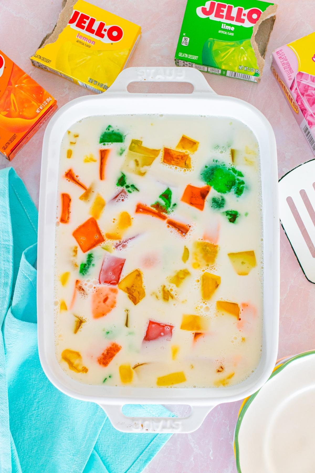 A white dish with colorful Jello cubes in creamy milk, surrounded by Jello boxes and a blue napkin.