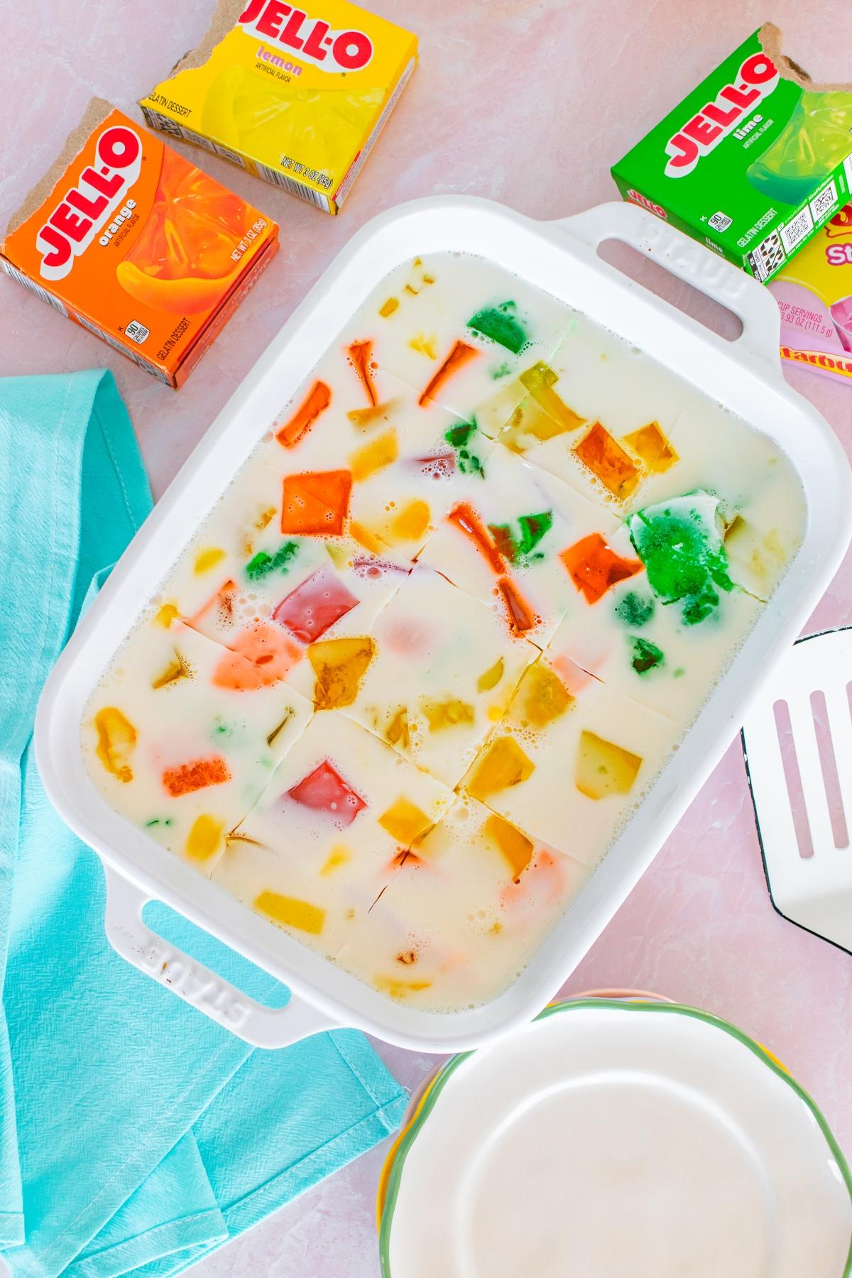 A white dish with colorful jello cubes in creamy liquid, surrounded by Jell-O boxes and a blue napkin.
